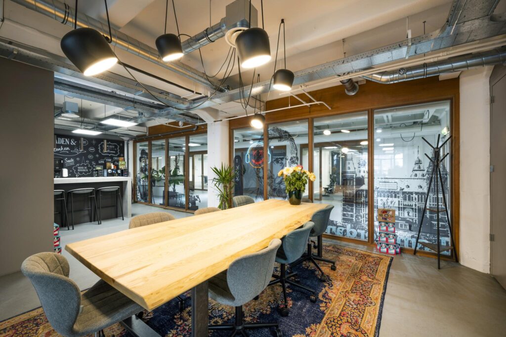 Modern meeting room on Van Diemenstraat with a large wooden table, industrial lighting, and glass walls featuring artistic Amsterdam-themed designs.